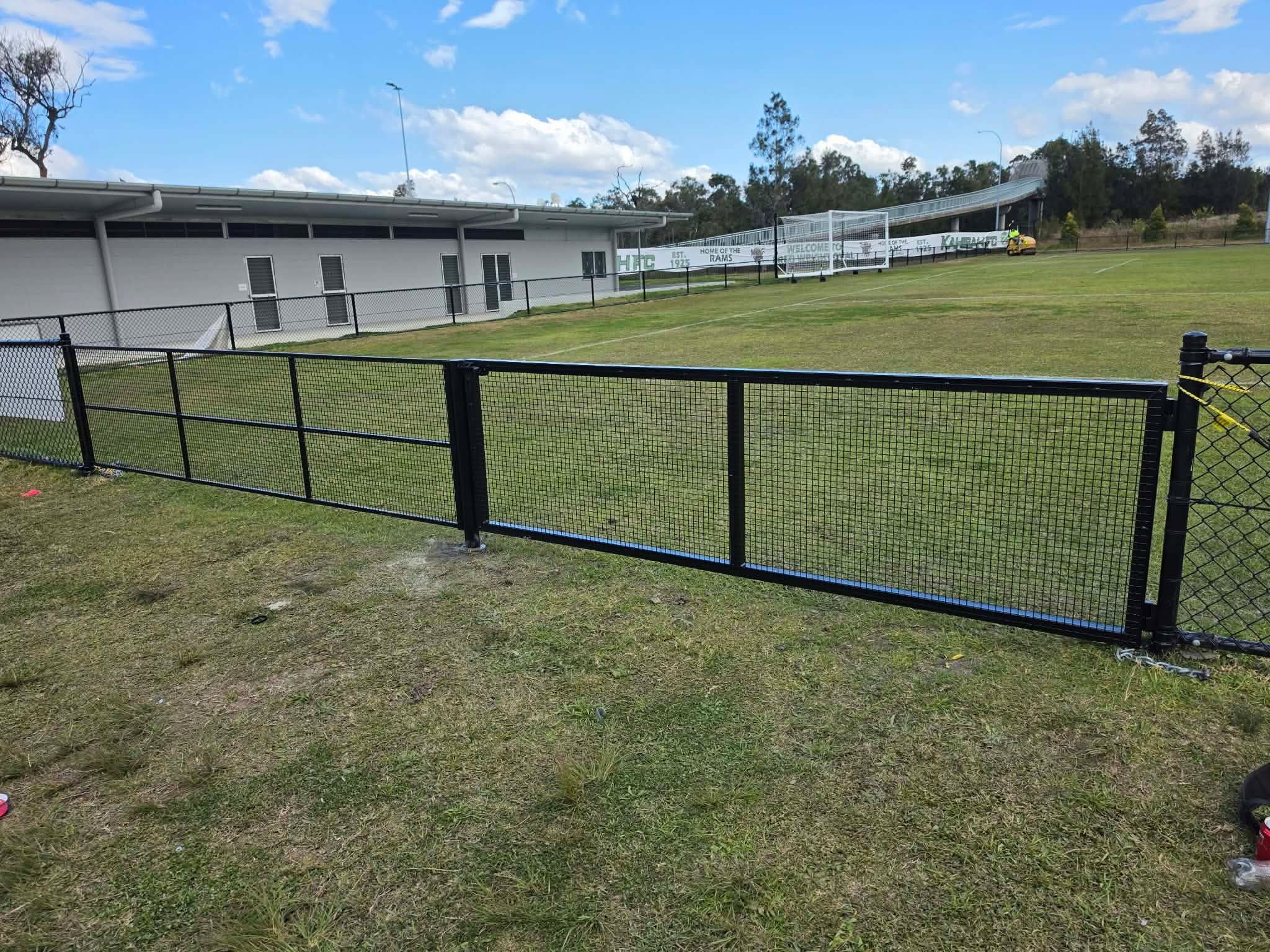 Black framed mesh gate set into a fence line beside a grassed sports field